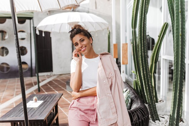 Pretty tanned woman in pink and white outfit leans on chair under umbrella in street cafe