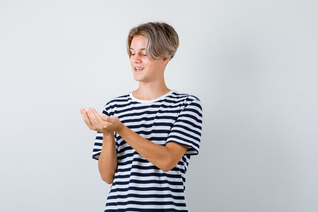 Pretty teen boy looking at his cupped hands in striped t-shirt and looking cheerful , front view.