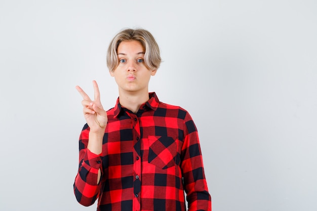 Pretty teen boy showing peace gesture, pouting lips in checked shirt and looking amazed , front view.
