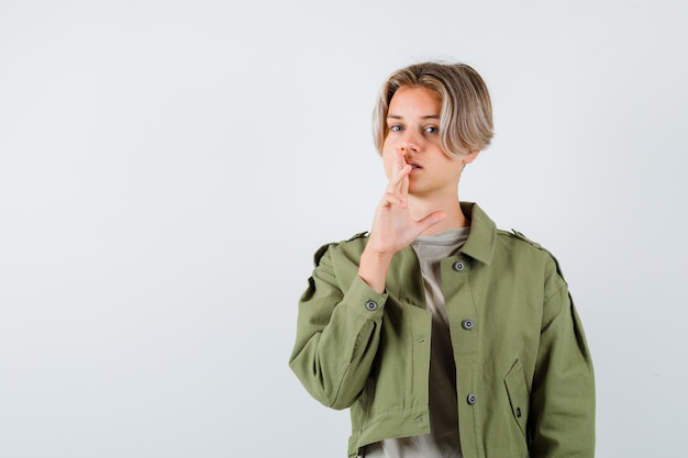 Pretty teen boy showing silence gesture in green jacket and looking sensible