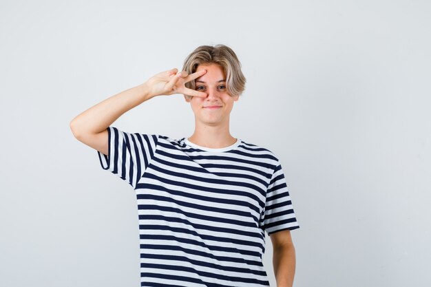 Pretty teen boy in striped t-shirt showing V-sign near eye and looking cheerful , front view.