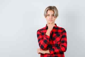 Free photo pretty teen boy with hand on chin in checked shirt and looking thoughtful. front view.