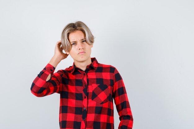 Pretty teen boy with hand behind ear, overhearing secret in checked shirt and looking confused , front view.