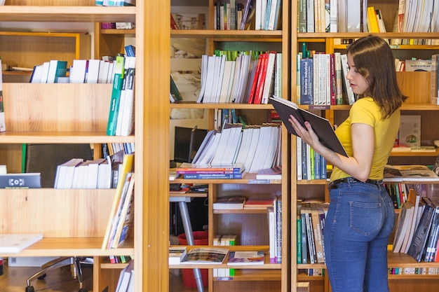 Ragazza abbastanza teenager che esamina libro vicino allo scaffale per libri