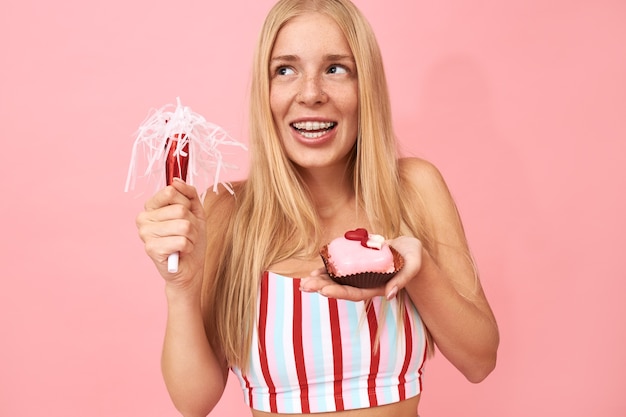 Pretty teen girl with straight hair and braces on her teeth celebrating birthday, posing isolated with party blower and sweet dessert, making wish, having dreamy joyful facial expression