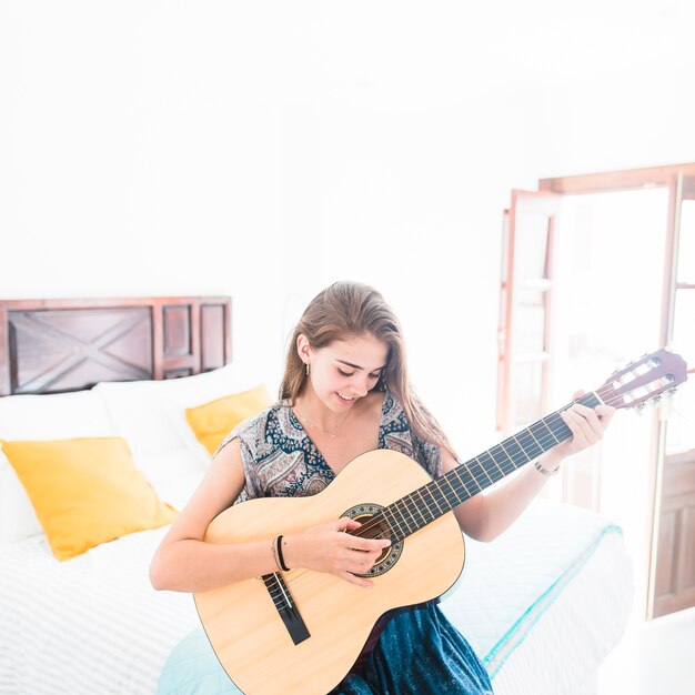 Довольно девочка-подросток, сидя на кровати, играя на гитаре