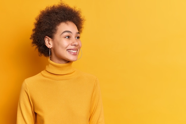 Pretty teenage girl with Afro hair looks gladfully aside smiles broadly being amused by someone dressed in casual turtleneck.