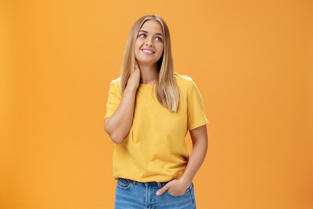 かわいくて優しいかわいいヨーロッパの女性の独立したフリーランサーは、黄色のTシャツで首に触れてため息をつき、オレンジ色の背景の上にポーズをとって、心地よい笑顔で右上隅を夢見ています。