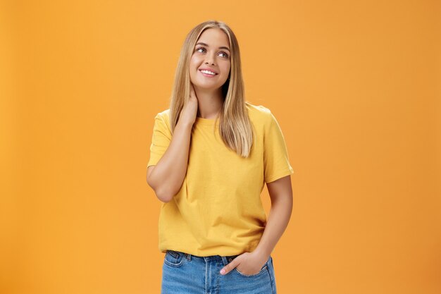Pretty and tender cute european female independent freelancer in yellow t-shirt sighing touching neck and gazing dreamy at upper right corner with pleasant smile, posing over orange background.
