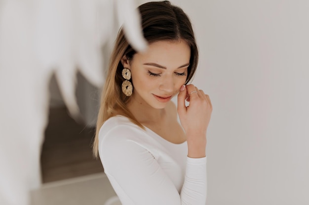 Pretty tenseness woman with nude make up wearing golden earrings is touching her cheek and smiling with closed eyes over isolated background