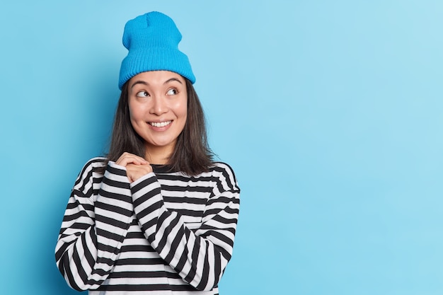 Pretty thoughtful asian woman keeps hands together looks aside with smile wears hat and striped jumper