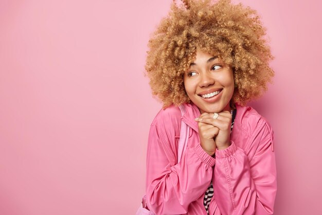 Pretty thoughtful woman has glad expression keeps hands under chin smiles toothily dressed in jacket focused away isolated over pink background with blank space for your promotional content