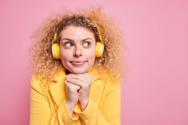 Pretty thoughtful woman keeps hands under chin being deep in thoughts has dreamy expression while listening favoriye lyrics songs via headphones wears yellow jacket poses against pink wall