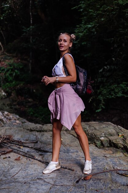 Pretty tourist in white crop top with backpack stands on rock in forest