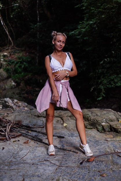 Pretty tourist in white crop top with backpack stands on rock in forest