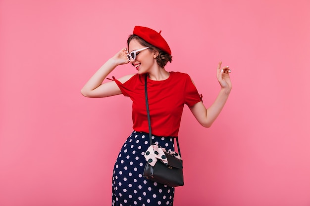 Pretty trendy woman in beret touching her sunglasses. Indoor photo of jocund curly brunette girl in french outfit.