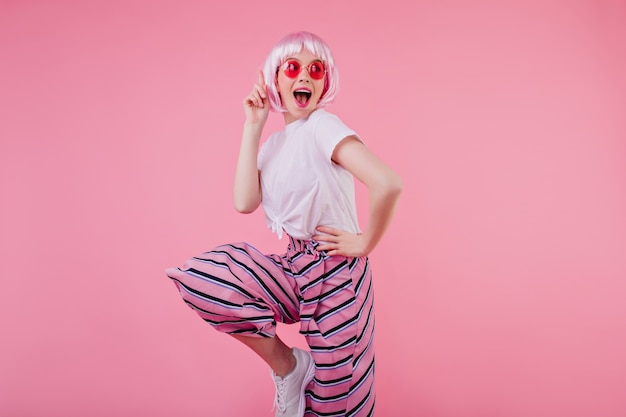 Pretty white girl in shiny peruke posing in funny pink sunglasses. Indoor photo of graceful lady in trendy pants enjoying photoshoot and smiling