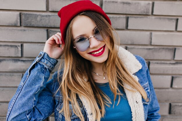 추운 봄 날에 재미 밝은 미소로 예쁜 백인 소녀. 즐거운 금발 여자의 야외 초상화는 파란색 안경과 빨간 모자를 착용합니다.