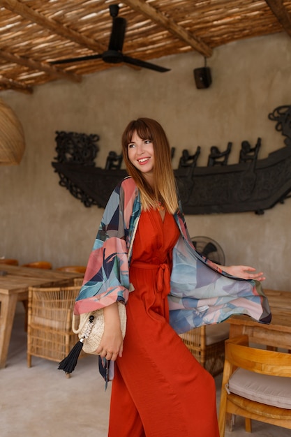 Pretty white woman in stylish summer bohemain autfit posing in tropical cafe.