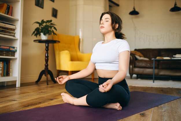 Pretty with chubby cheeks sitting on yoga mat with legs folded, closing eyes, taking deep breath, practicing meditation, searching for inner peace and harmony.