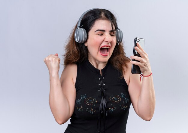 Pretty woman in black blouse holding phone listening to music with headphones and dancing 