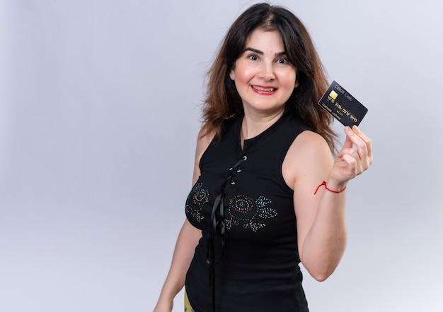 Красивая женщина в черной блузке показывает кредитную карту и радостно смотрит в камеру