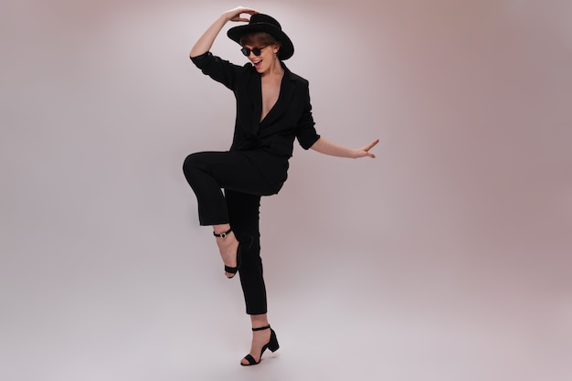 Pretty woman in black outfit and hat moves on white background. Charming lady in dark jacket and pants dances and jumps on isolated