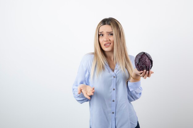 Pretty woman in blue outfit posing with single cabbage on white wall.