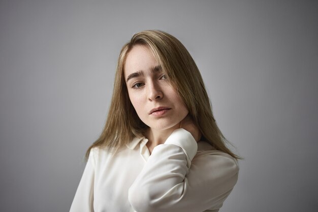 Pretty woman of Caucasian appearance having serious facial expression, rubbing her neck. Beautiful young female with natural make up and loose fair hair, posing in studio with hand at her face