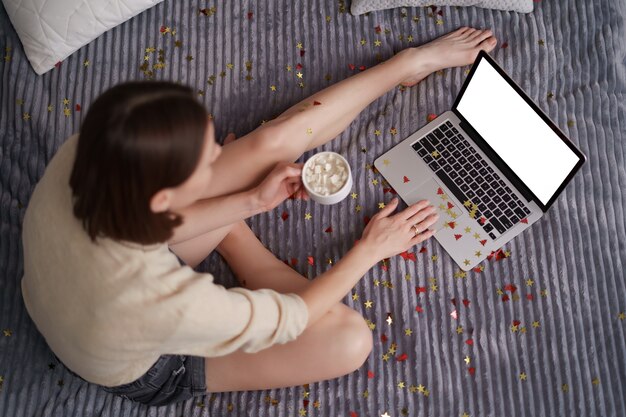 Pretty woman celebrating with her family and friends online using laptop, drinking coffee with confetti on bed
