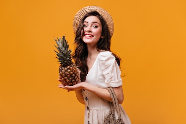 Pretty woman in abito di cotone esamina la macchina fotografica con un sorriso e pose con ananas su sfondo isolato.