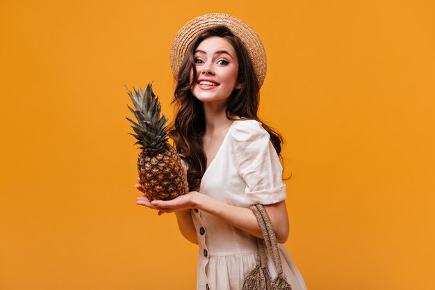 Pretty woman in cotton dress looks into camera with smile and poses with pineapple on isolated background.