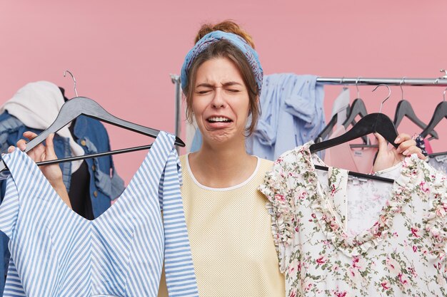 Pretty woman crying while standing in cloakroom, holding two fashionable dresses of high price, having no money to buy them. Upset, sorrorful female can`t find something suitable for herself