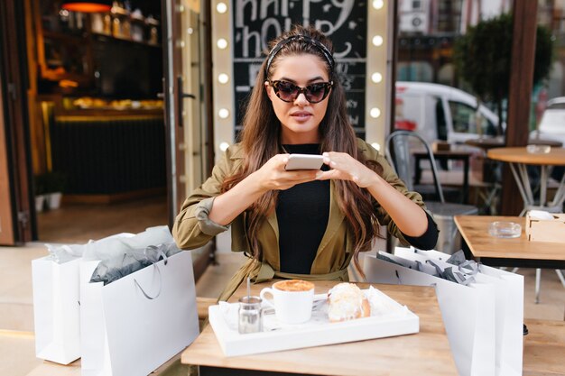 Pretty woman in dark sunglasses sitting in outdoor cafe beside bags from boutique