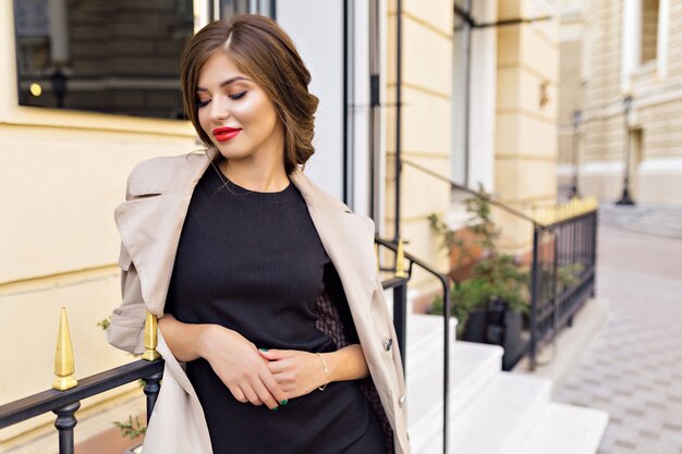 Pretty woman dressed in black dress and beige trench with stylish hairstyle and red lips at the street