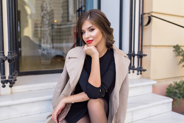 Foto gratuita bella donna vestita in abito nero e trench beige con acconciatura alla moda e labbra rosse in strada
