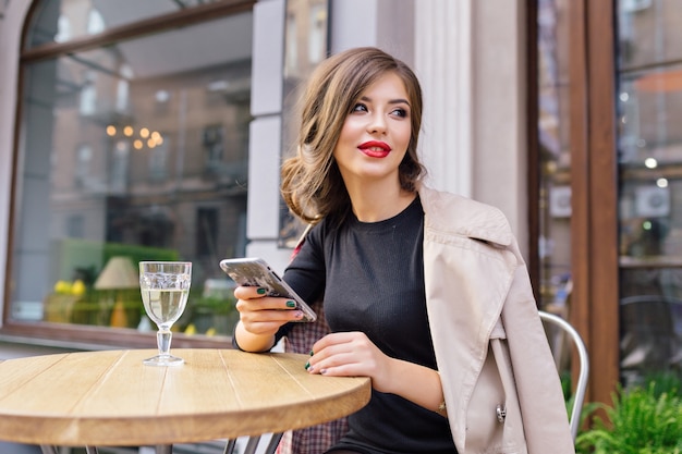 Pretty woman dressed in black dress and beige trench with stylish hairstyle and red lips on a terrace