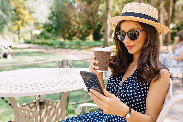 Pretty woman dressed in dress, summer hat and sunglasses is sitting in summer cafeteria and rests. She drinks coffee and looking in her phone with light smile. Beautiful portrait. Place for text.