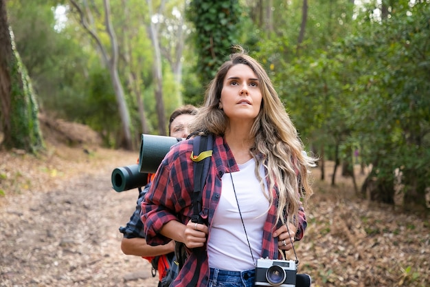 Pretty woman enjoying view and hiking. Couple tourists walking in forest together. Young Caucasian hikers or traveler with backpacks trekking together. Tourism, adventure and summer vacation concept