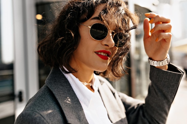 Pretty woman in grey jacket and white shirt in sunglasses with red lipstick is touching hair and posing near her house Beautiful lady in grey oversize jacket and pants walks outside