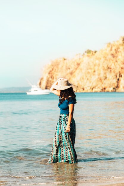 Милая женщина в шляпе стоя в прибрежной волне на пляже