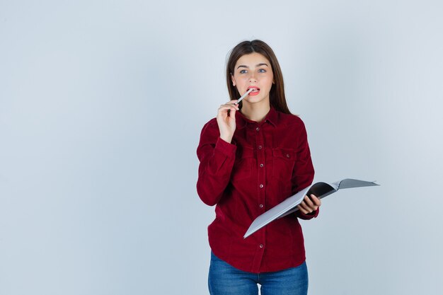 Pretty woman holding folder, keeping pen between teeth in burgundy blouse and looking preoccupied