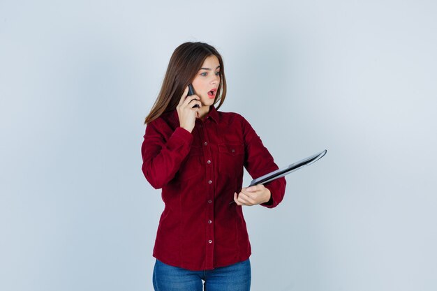 Pretty woman holding folder, talking on smartphone in burgundy blouse and looking shocked