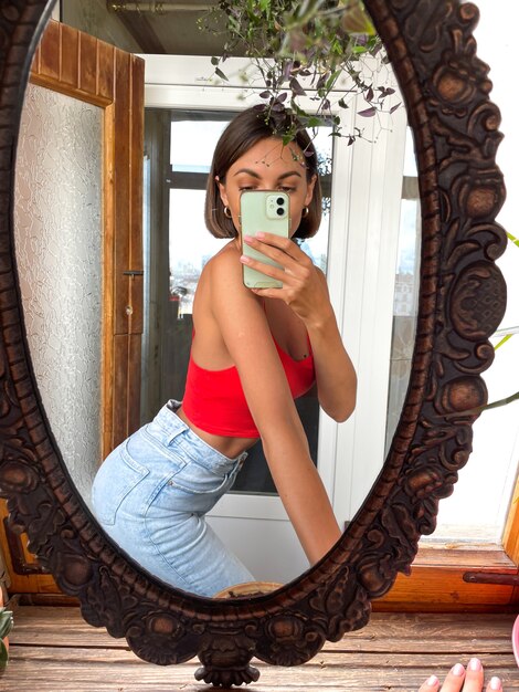 집에서 예쁜 여자는 청바지와 빨간 탑을 입고 소셜 미디어의 이야기와 게시물을 위해 휴대 전화의 거울에 사진 셀카를 찍습니다.