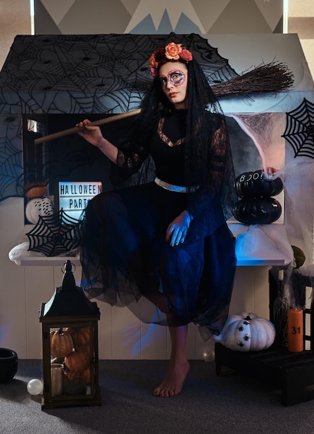 Pretty woman is celebrating halloween in black dress and unusual makeup while holding broom.