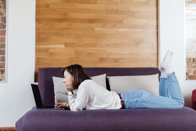 きれいな女性は自宅の木製の壁の前に暗いベッドの上のラップトップコンピューターに取り組んでいます