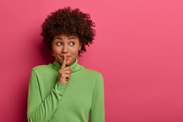 Pretty woman makes shush, asks not be loud, has secret expression, presses index finger over lips, has mysterious expression, wears green jumper