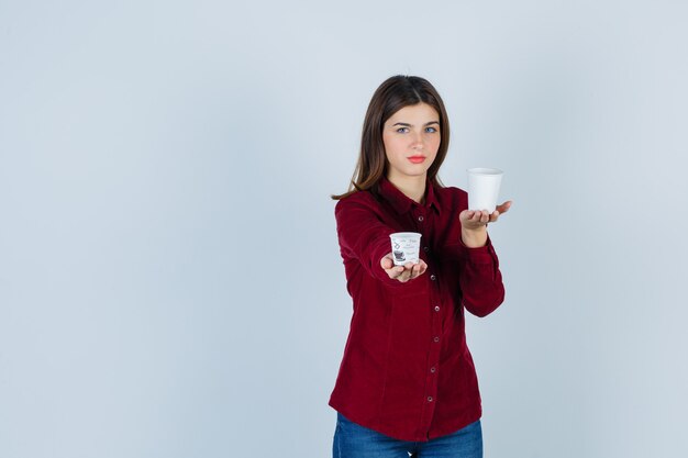 Симпатичная женщина в бордовой блузке с двумя пластиковыми чашками кофе выглядит уверенно.