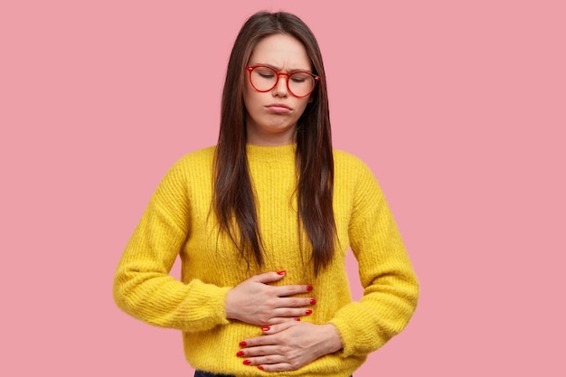 Бесплатное фото Симпатичная женщина давит руками на живот, чувствует боль и дискомфорт, имеет проблемы со здоровьем и дискомфорт при приеме пищи, носит желтый свитер, позирует на розовом фоне