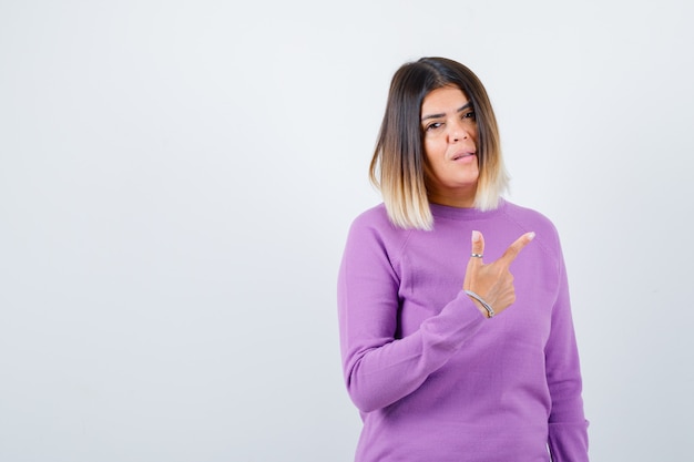 Pretty woman in purple sweater pointing at upper right corner and looking indecisive , front view.